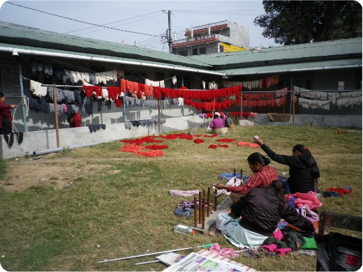 屋外で座り作業をしている人と周りには赤い布が干されている写真